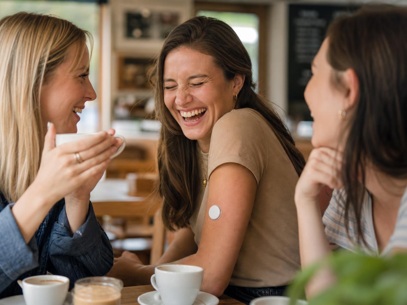 Three friends laughing over coffee, one wearing a glucose sensor on her upper arm