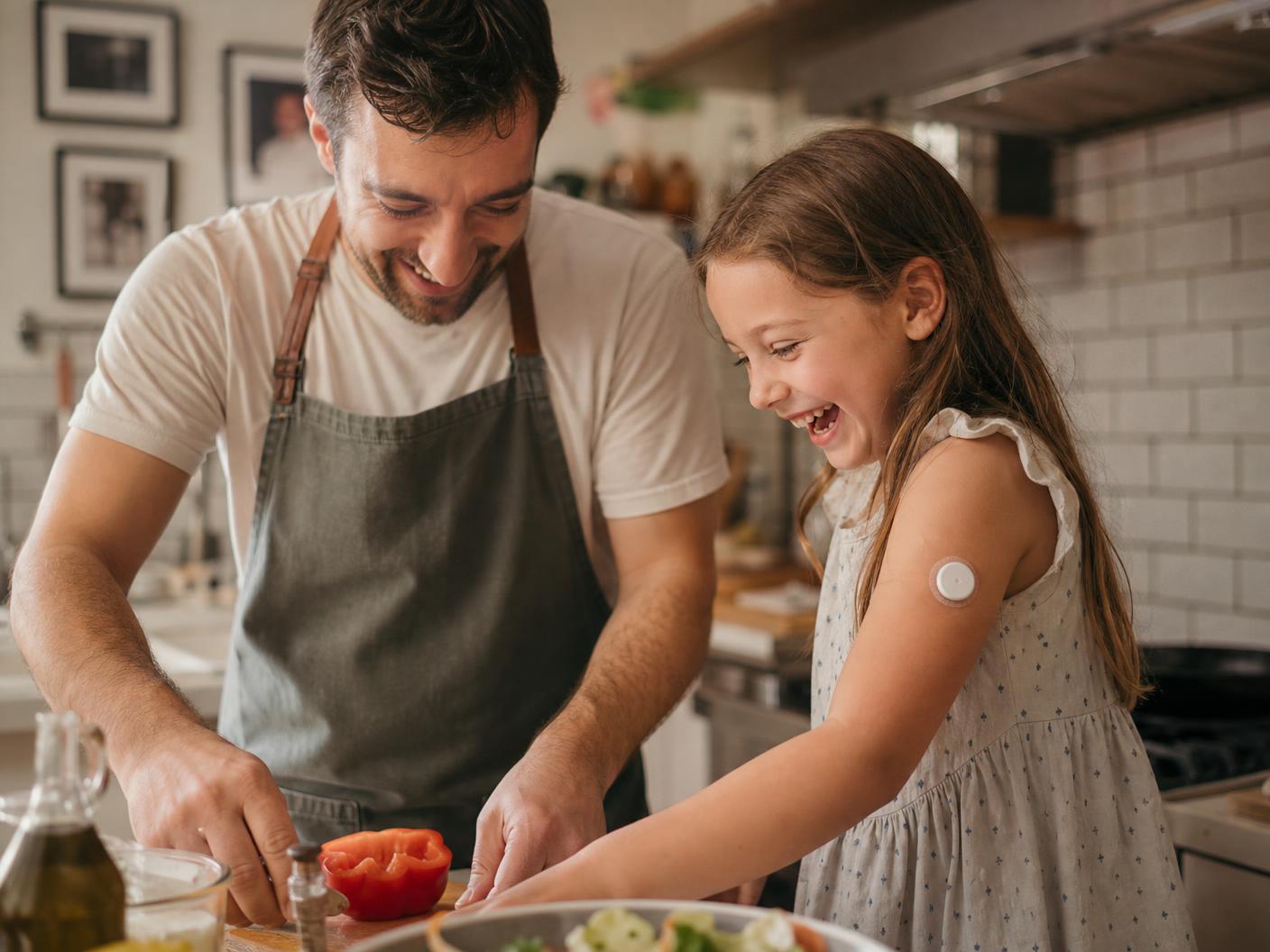 A father and his daughter, who wears a continuous glucose sensor on her arm, cooking dinner together in a sunlit kitchen